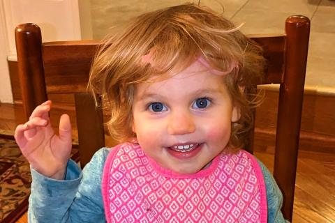 A toddler, Leah, smiles and waves at the camera.