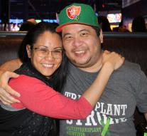 Angelo and a woman sit in a booth together, arms around each other, smiling at the camera.